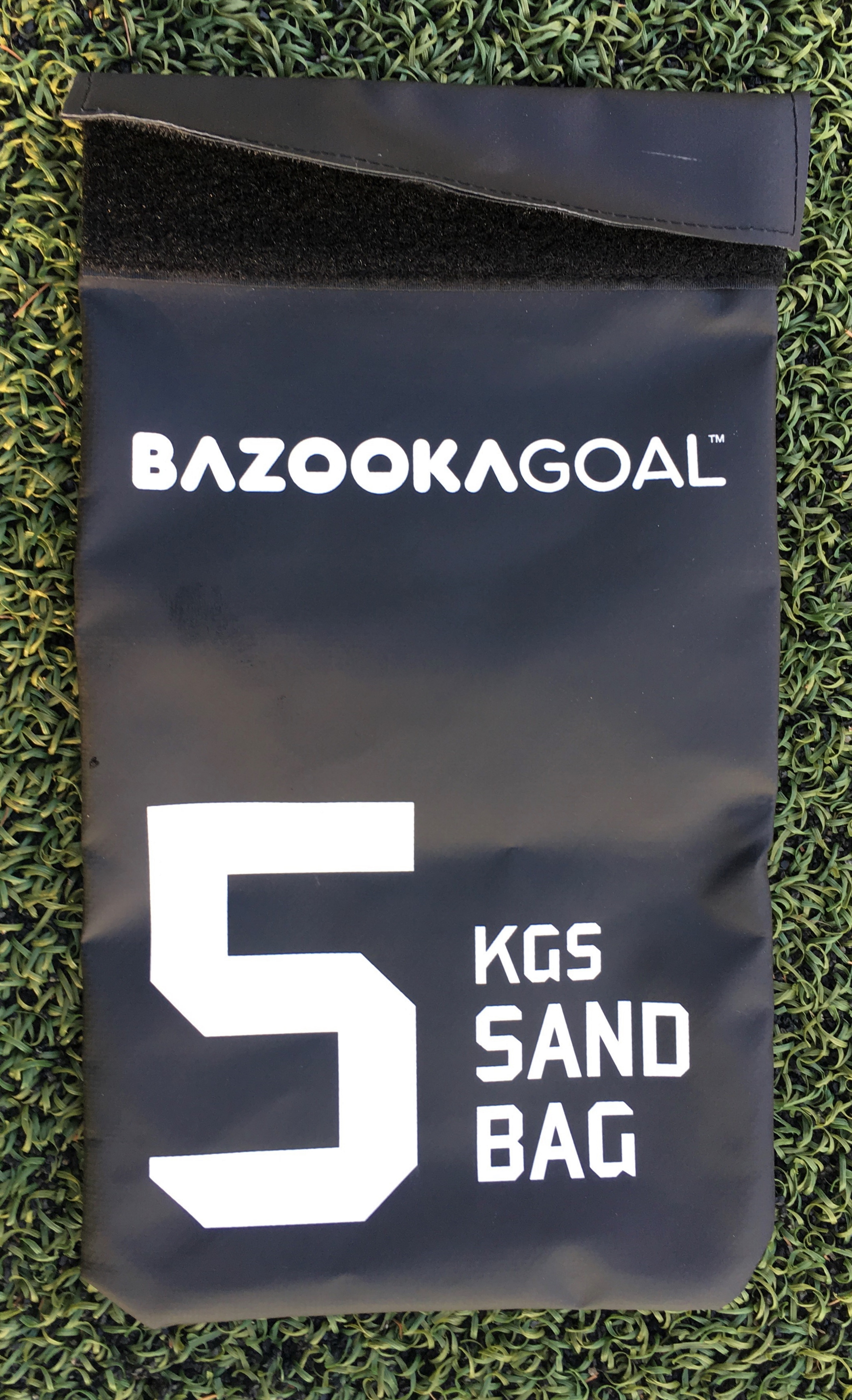 Ein schwarzer BazookaGoal Sandsack mit einem Gewicht von 5 kg, platziert auf einem Kunstrasen.